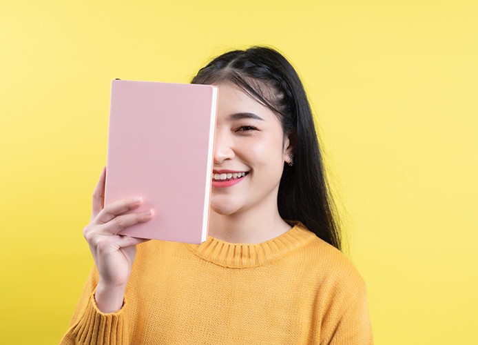 Meisje voor gele achtergrond houdt een boek voor haar gezicht en kijkt lachend in de camera