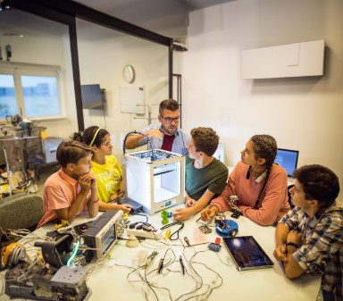 Groep jongeren staat rondom een 3D-printer en luistert aandachtig naar een volwassen man die iets uitlegt over de 3D-printer.