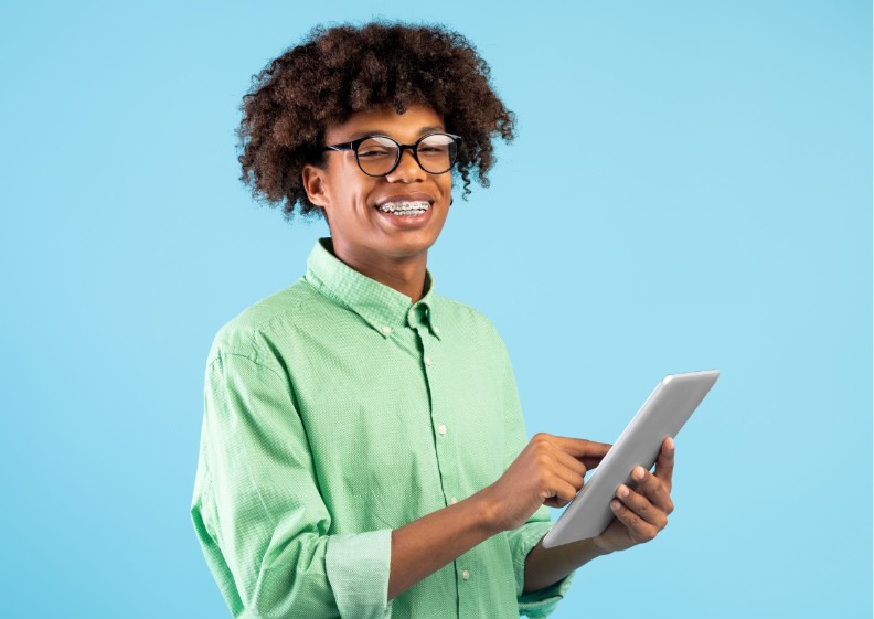 Tienerjongen met een bril en een beugel houdt een tablet vast en kijkt met een brede lach in de camera.