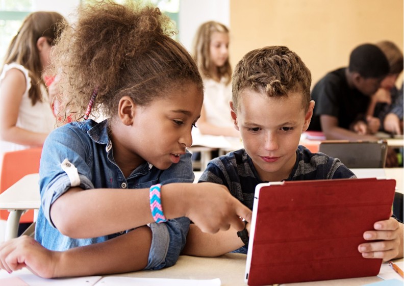 Een jongen heeft een tablet vast. Een meisje zit naast de jongen en wijst iets aan op de tablet.